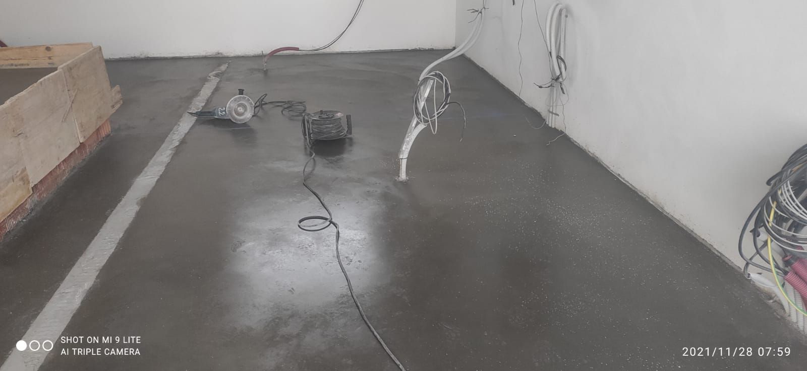 Habitación vacía con suelo oscuro y mojado, mangueras y equipos de limpieza cerca de paredes blancas.