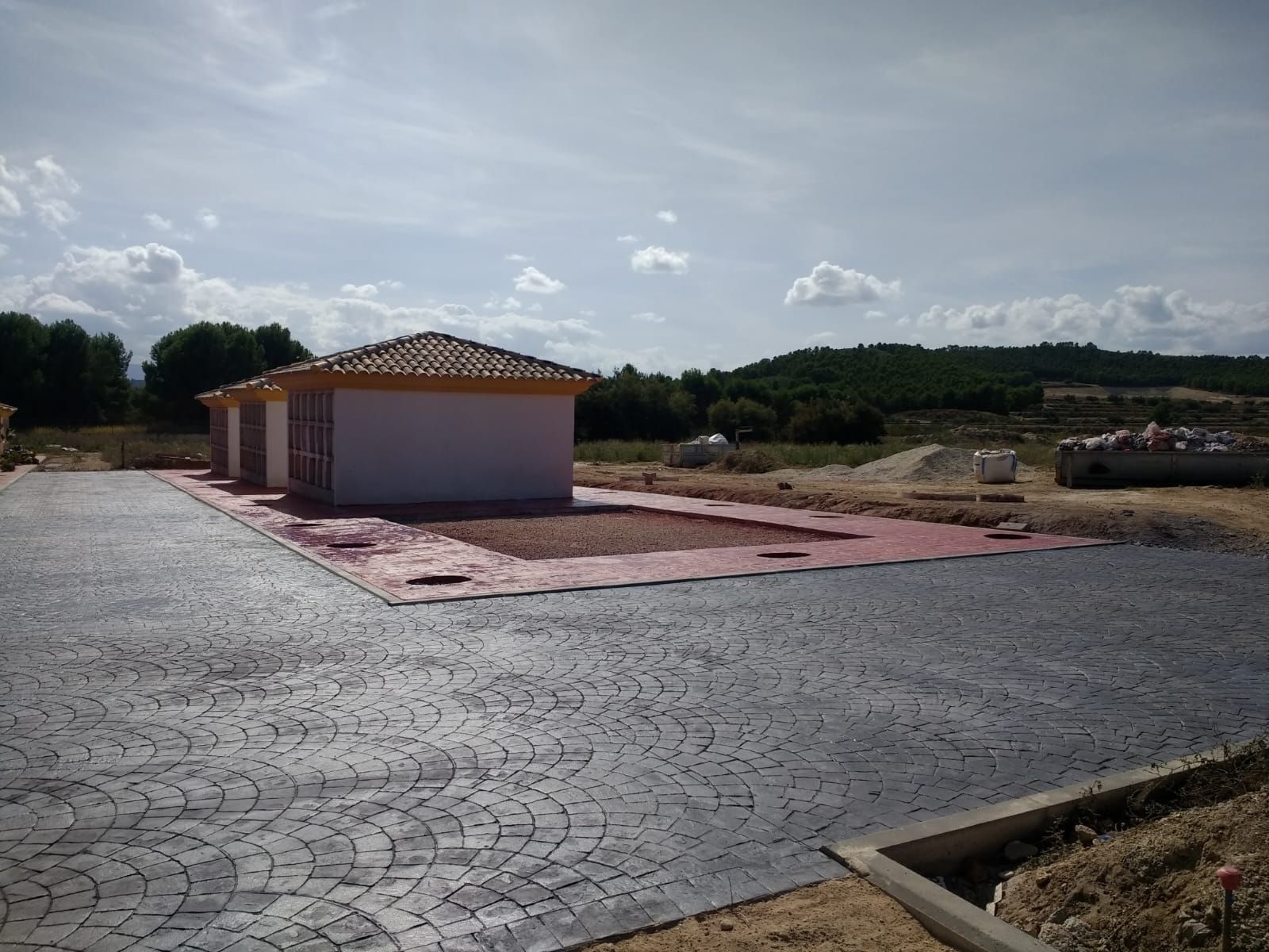 Pequeña casa con tejas en construcción sobre un terreno pavimentado, rodeada de tierra desnuda bajo un cielo nublado.