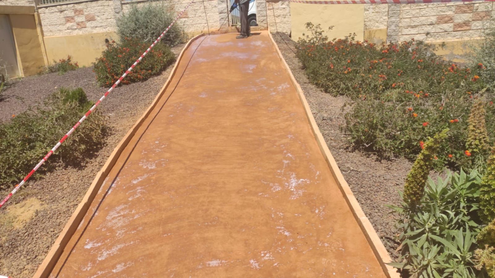 Pista de tenis de arcilla naranja con red, bordeada de plantas y un muro de piedra bajo un sol brillante.
