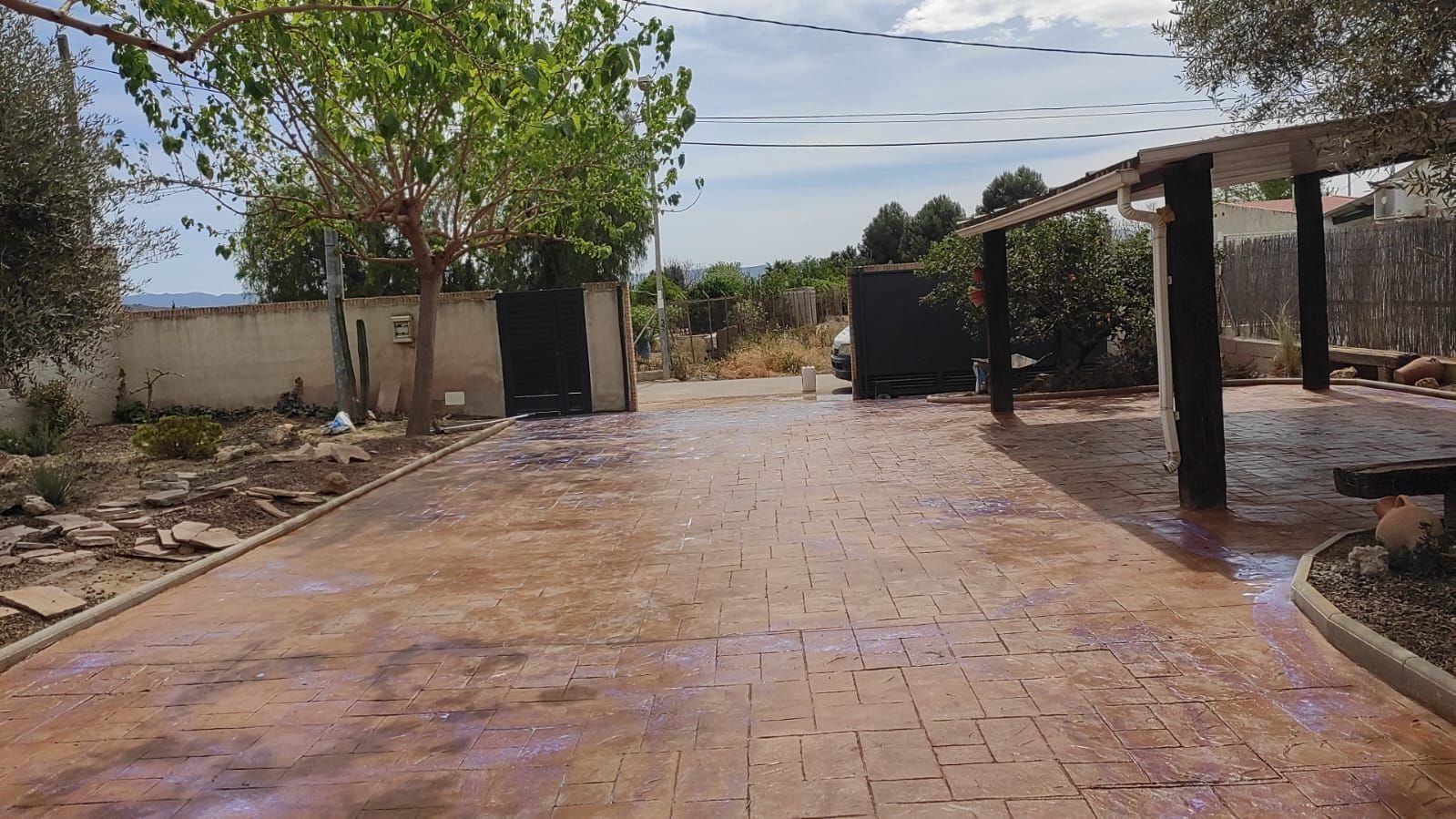 Un camino de ladrillos mojado con charcos, árboles y una puerta junto a una valla en un día soleado.
