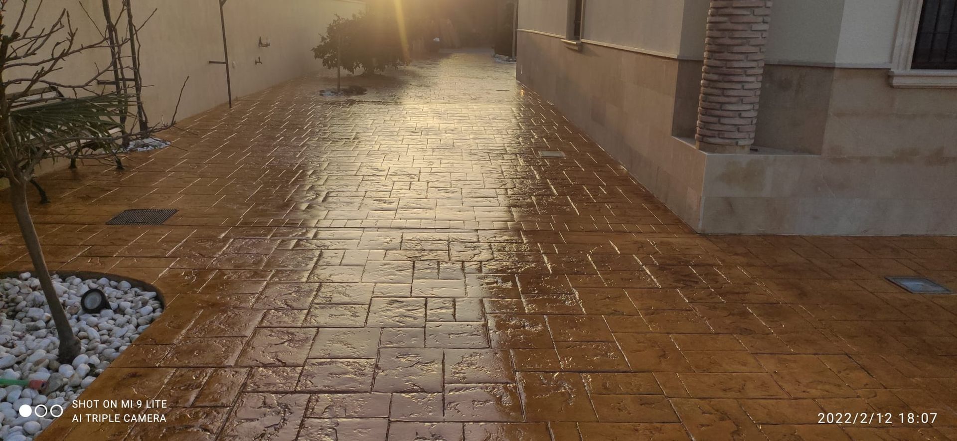 Un sendero lluvioso junto a un edificio al atardecer, con el pavimento de ladrillo mojado y los reflejos en el suelo.