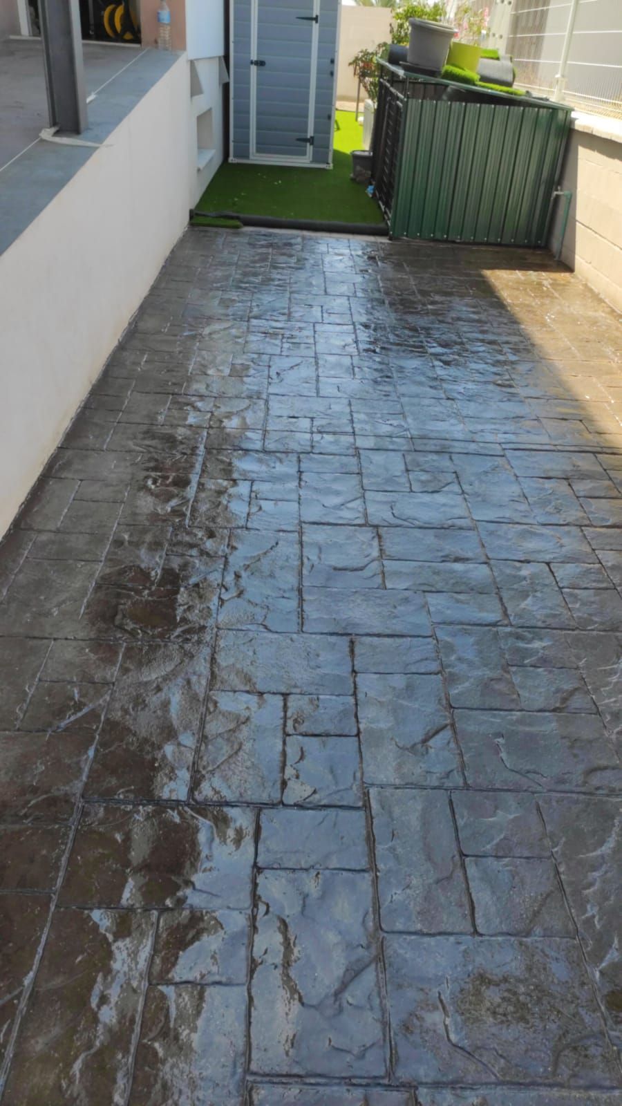 Un estrecho sendero de piedra mojada junto a un edificio conduce a una puerta y una valla verdes bañadas por el sol.