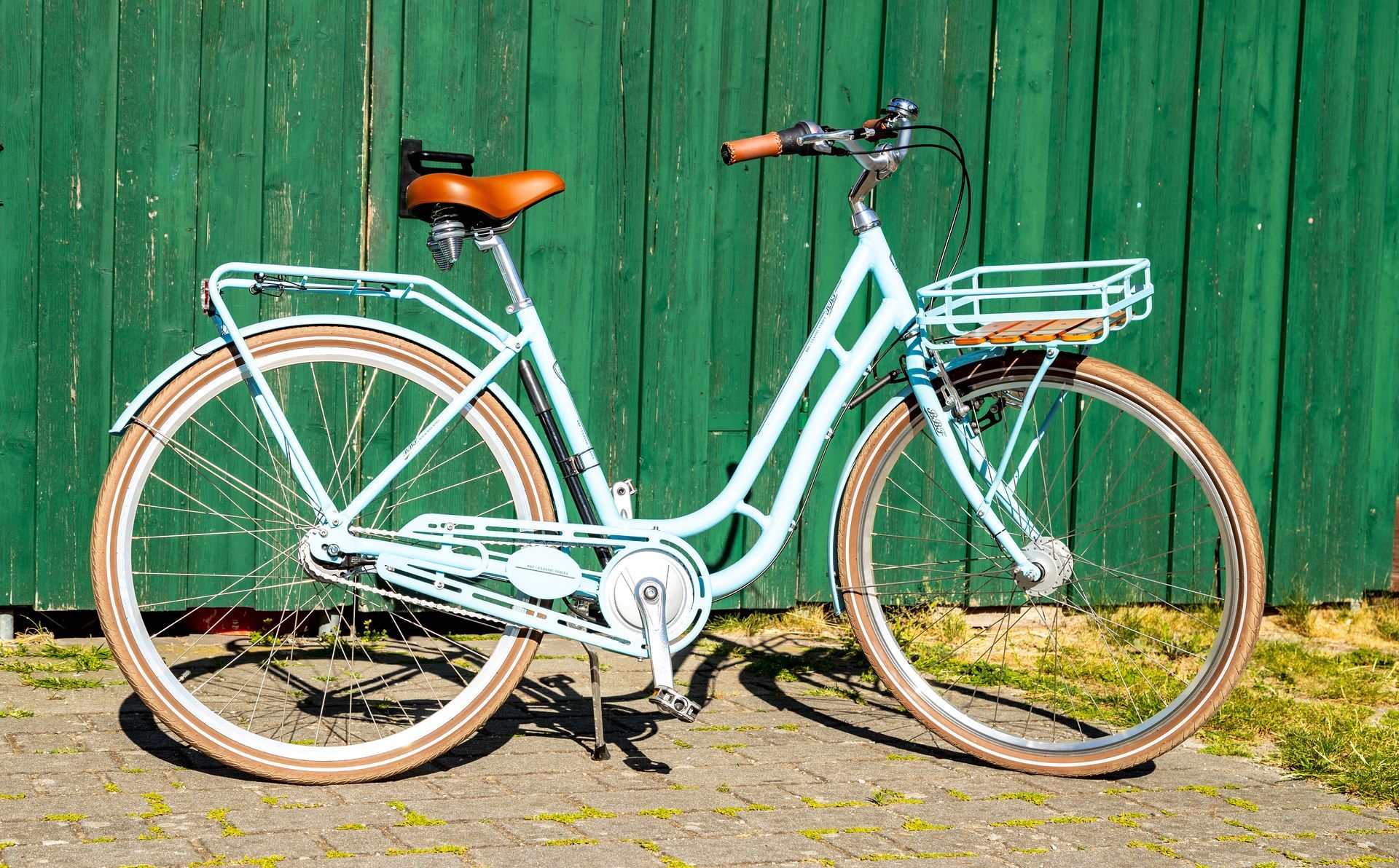 Hellblaues Fahrrad mit braunem Sattel und Korb vor einer grünen Holzwand.