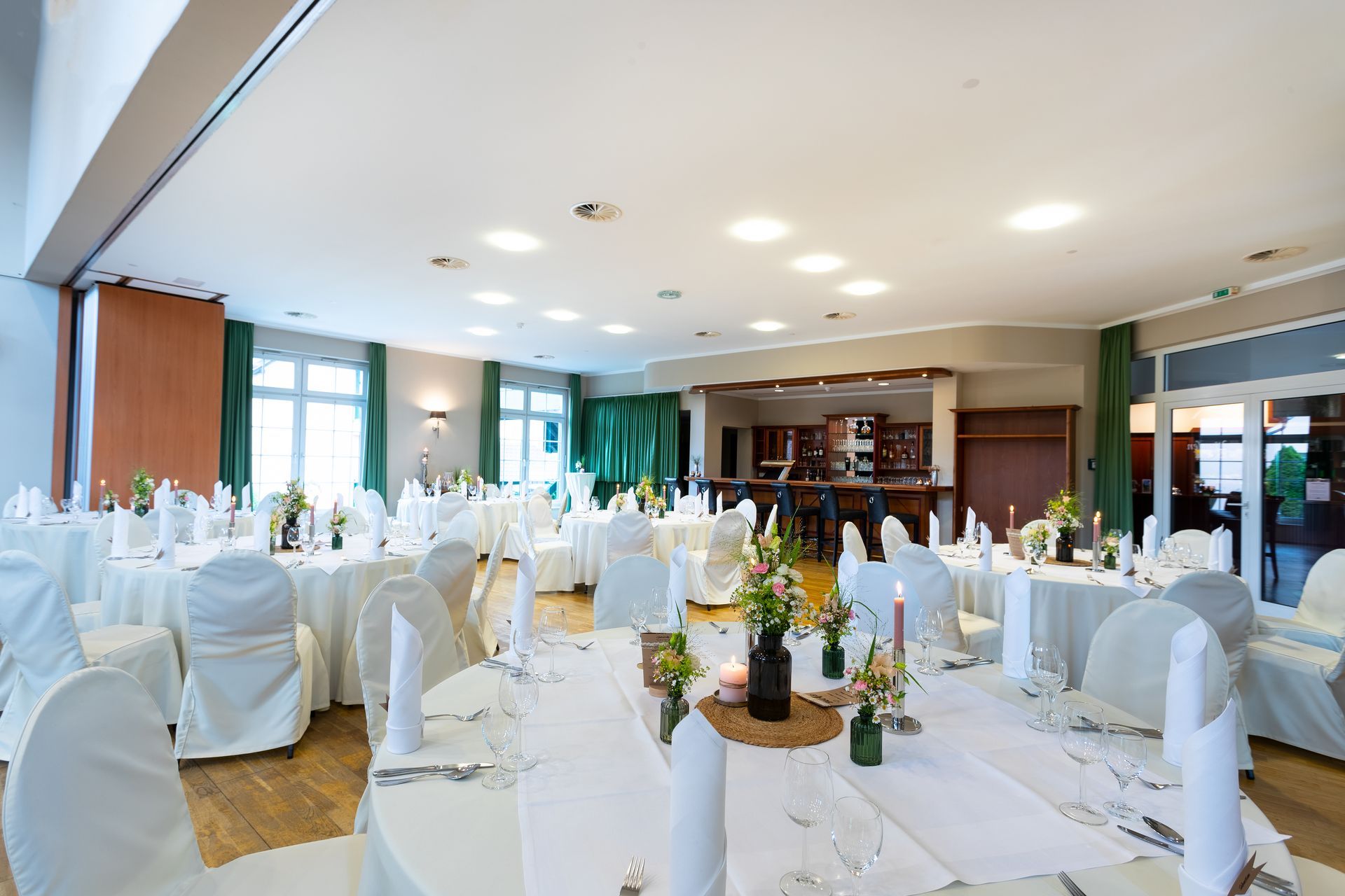 Hochzeitsempfangssaal mit runden Tischen, weißer Tischwäsche, Blumengestecken und einer Bar.