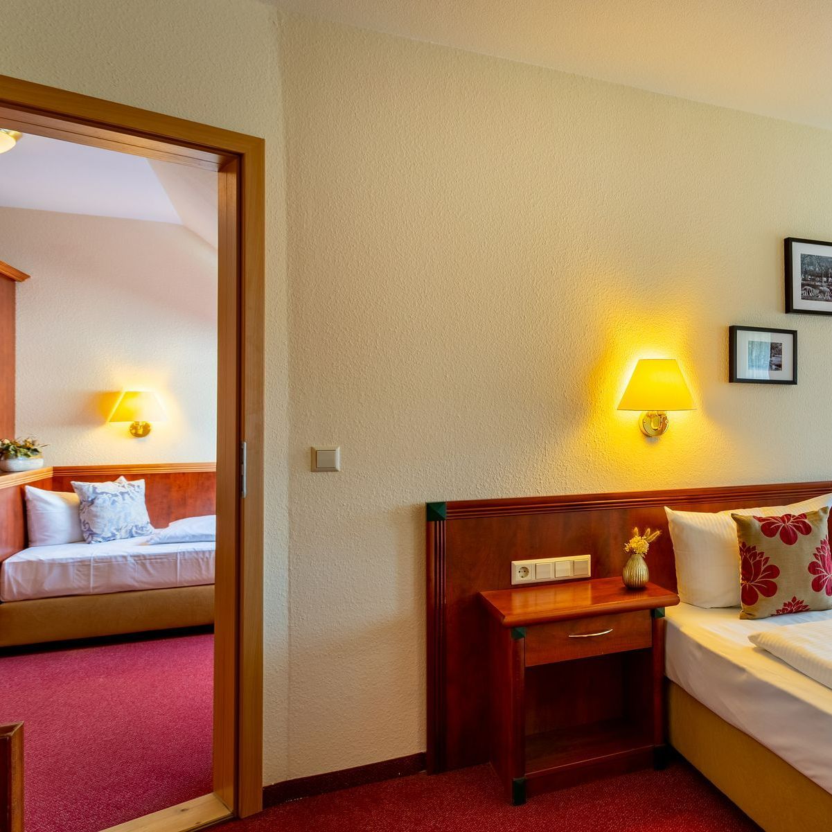 Hotelzimmereinrichtung mit Bett, Nachttisch und Blick in ein anderes Zimmer. Roter Teppich und warme Beleuchtung.