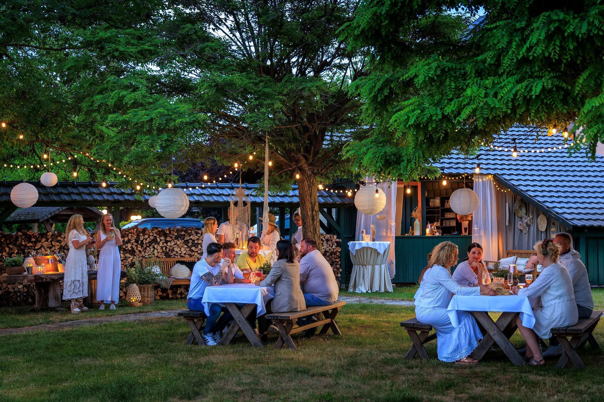 Menschen bei einem abendlichen Treffen im Freien, an Tischen sitzend, unter Bäumen mit Lichterketten und Papierlaternen.