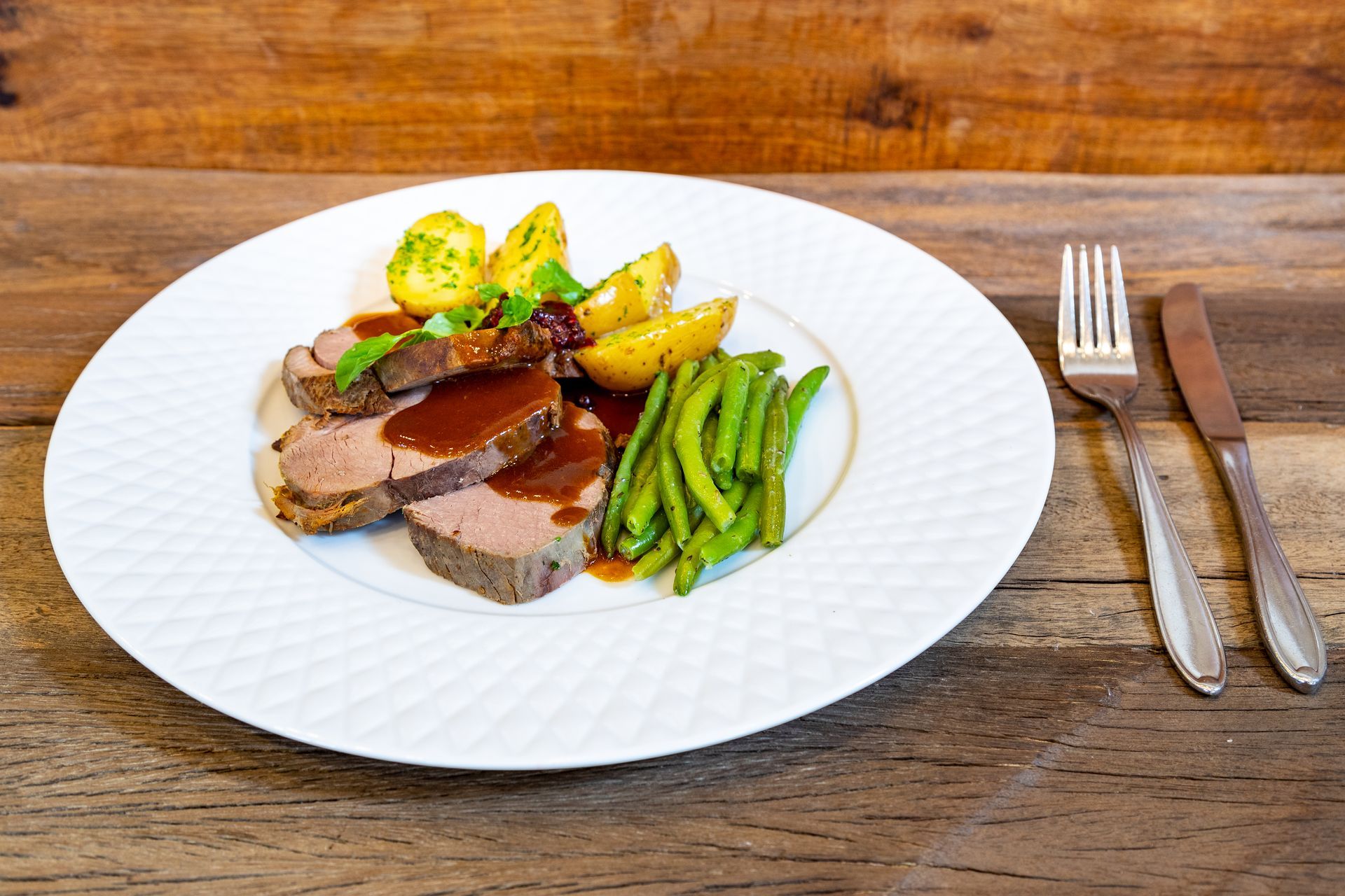 Roastbeefscheiben mit Soße, Kartoffeln und grünen Bohnen auf einem weißen Teller, mit Besteck.