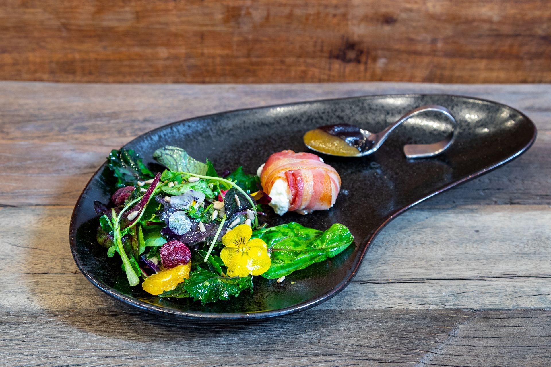 Vorspeisenteller: Salat mit Orangenscheiben, mit Speck umwickeltes Essen, kleiner Löffel mit Soße, auf dunklem Teller, Holzhintergrund.