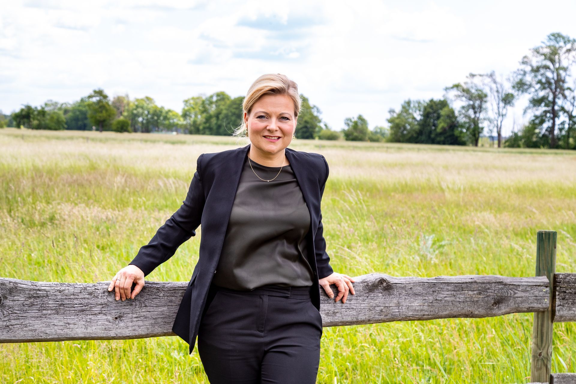 Frau im schwarzen Anzug, lächelnd an einem Holzzaun auf einem Feld gelehnt.