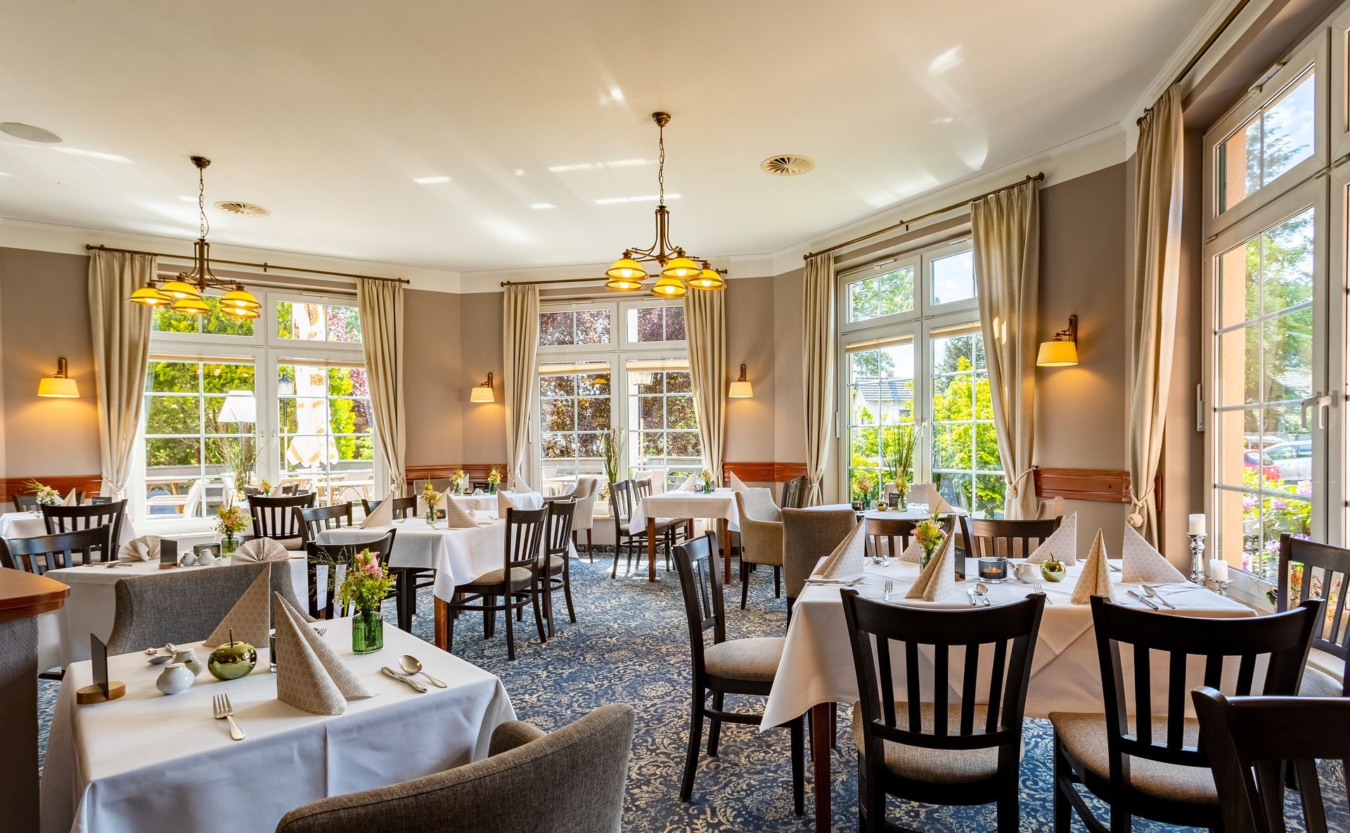 Restauranteinrichtung mit weißen Tischdecken, Holzstühlen, großen Fenstern mit Vorhängen und Blumendekorationen.
