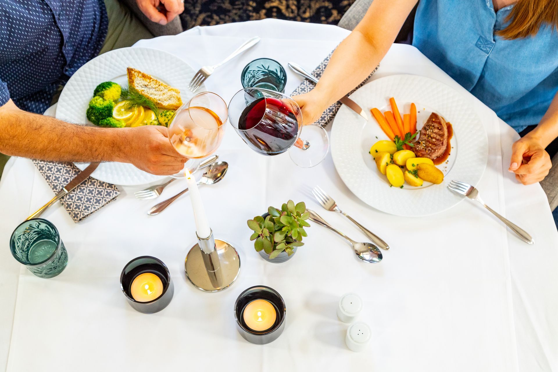 Ein Paar stößt mit Weingläsern über einem Esstisch mit Tellern voller Essen und Kerzen an.