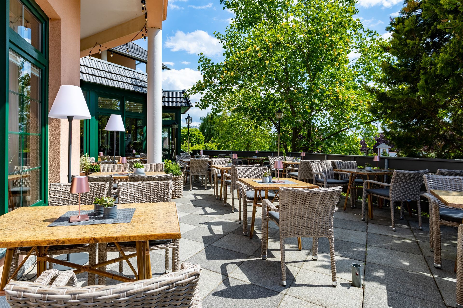 Außenterrasse eines Restaurants mit Tischen, Stühlen und grünen Bäumen; Sonnenlicht.