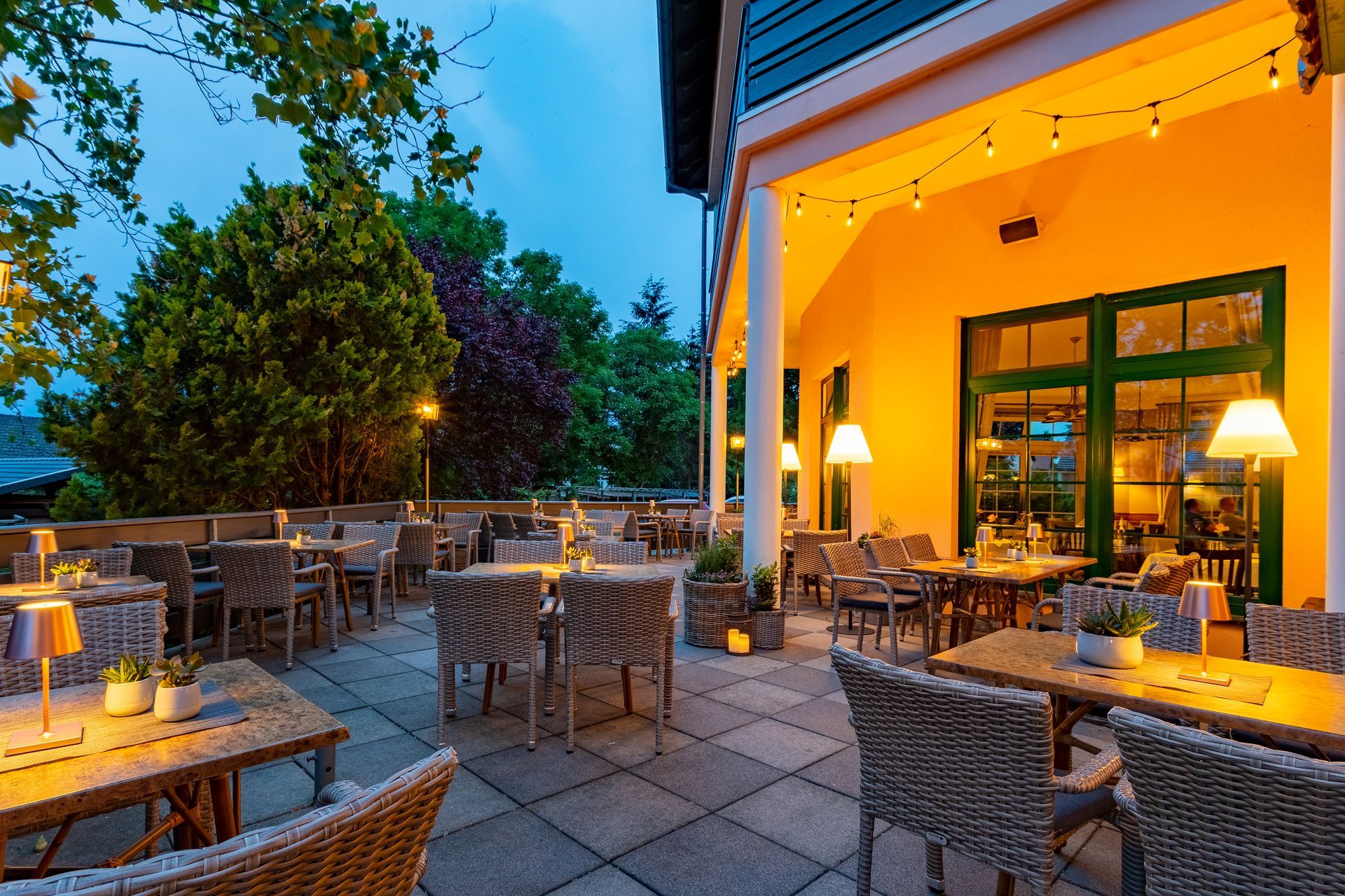Außenterrasse eines Restaurants mit Tischen, Stühlen und warmer Beleuchtung; Abendstimmung mit Bäumen und einem Gebäude.