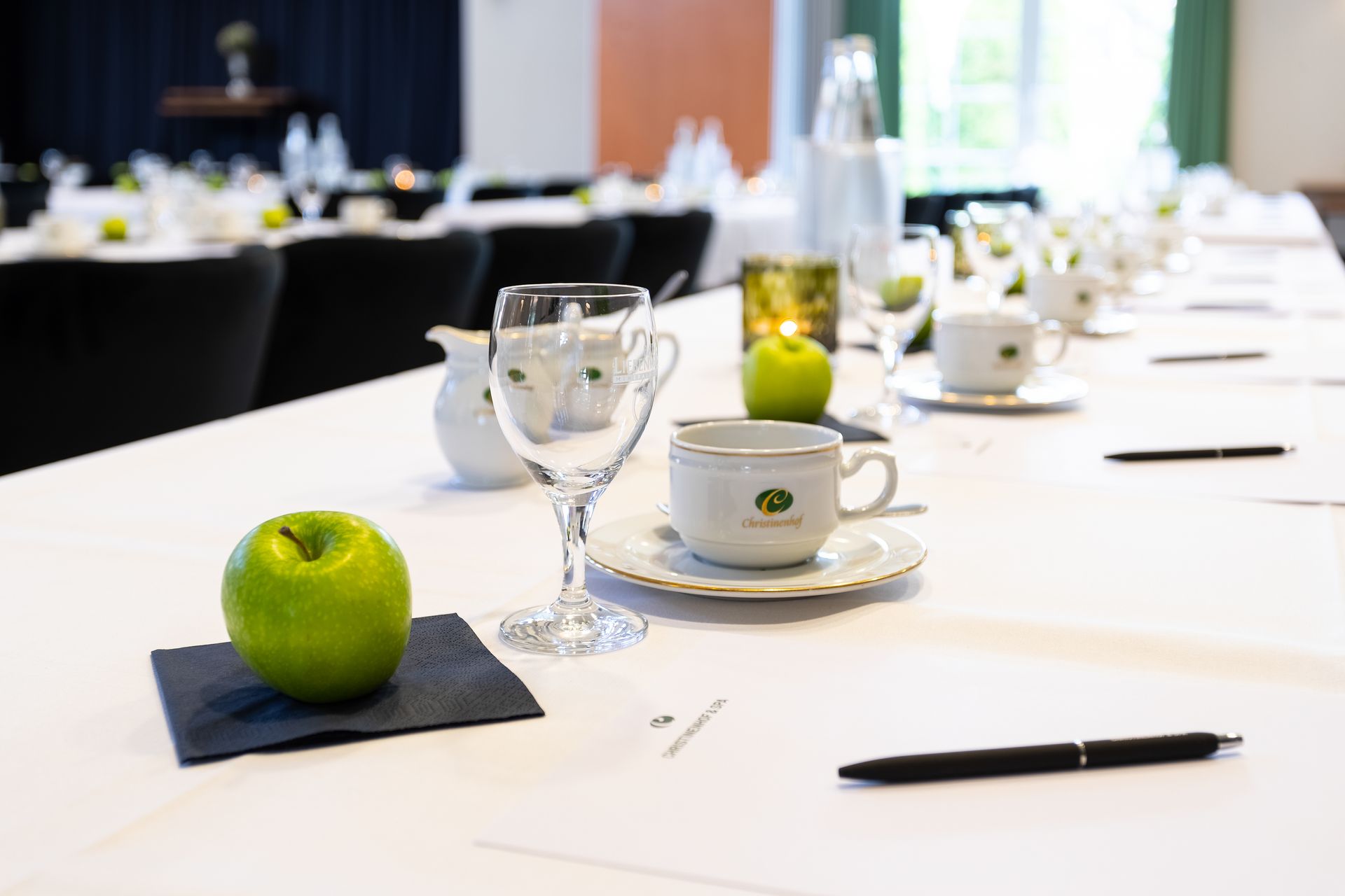 Langer Konferenztisch mit weißer Tischdecke, Äpfeln, Gläsern und Kaffeetassen.