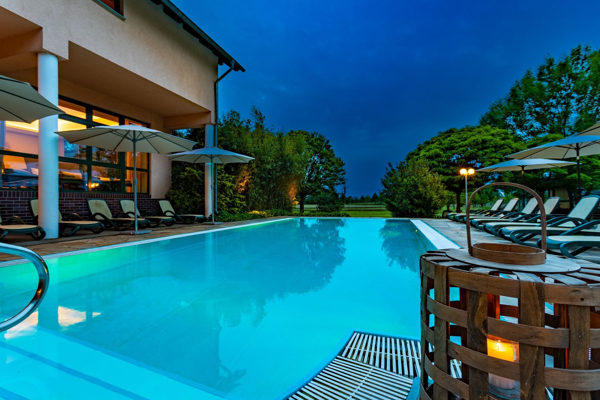 Pool in der Abenddämmerung mit Liegen, Sonnenschirmen und Gebäude; grün beleuchtetes Wasser, Bäume und leuchtende Laterne.