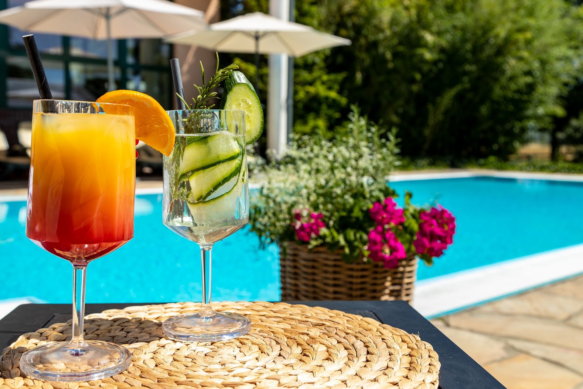 Zwei Cocktails an einem Pool mit Sonnenschirmen und Blumen.