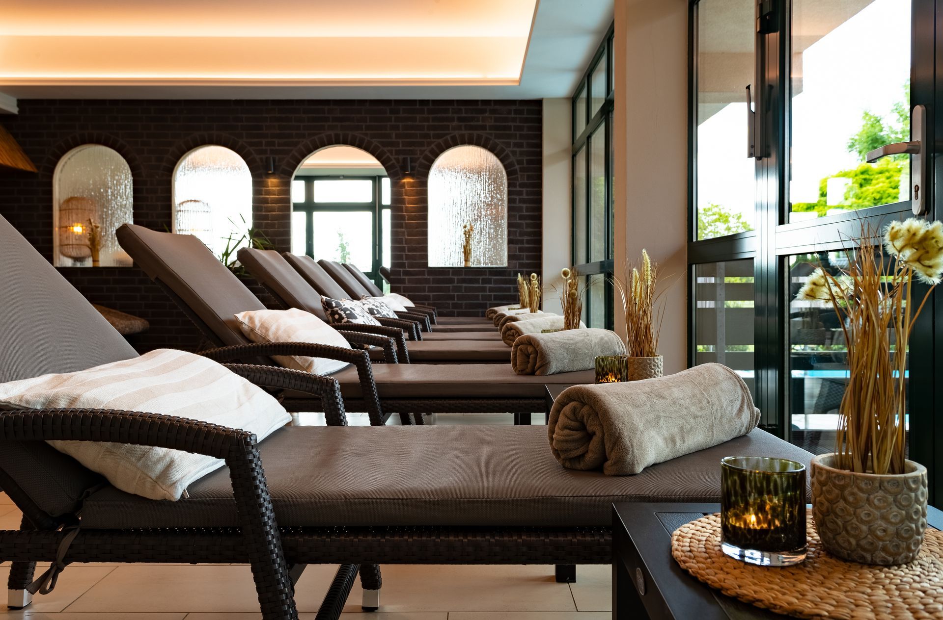 Spa-Lounge mit braunen Chaiselongues, Handtüchern und einer Ziegelwand mit Bogenfenstern.
