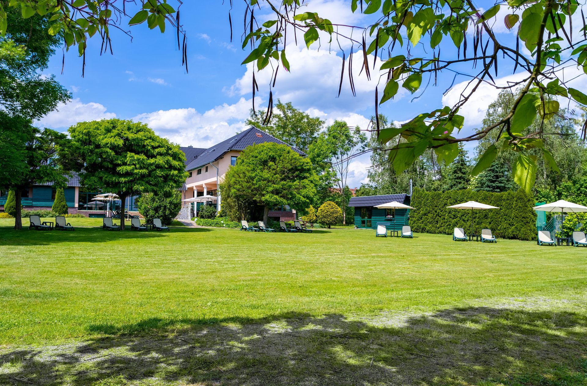 Üppiger grüner Rasen mit einem großen Haus, Bäumen, Sonnenschirmen und kleinen Gebäuden an einem sonnigen Tag.