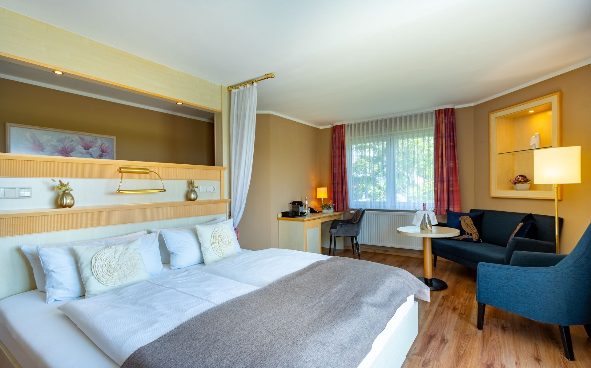 Hotelzimmer mit großem Bett, Schreibtisch, Couch und Fenster. Sanfte Farben, Holzakzente.