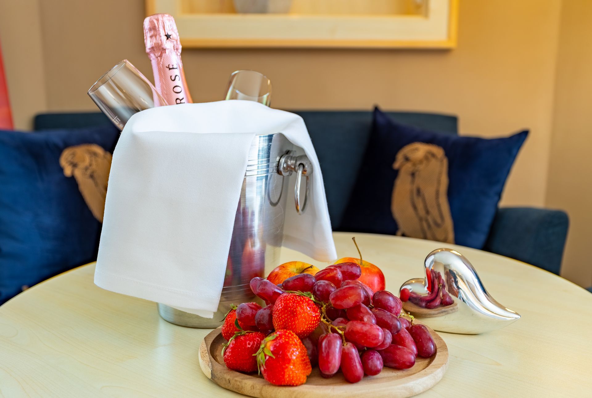 Ein Tisch mit einem Sektkühler, Obst und einem dekorativen Silberstück vor einem Sofa.