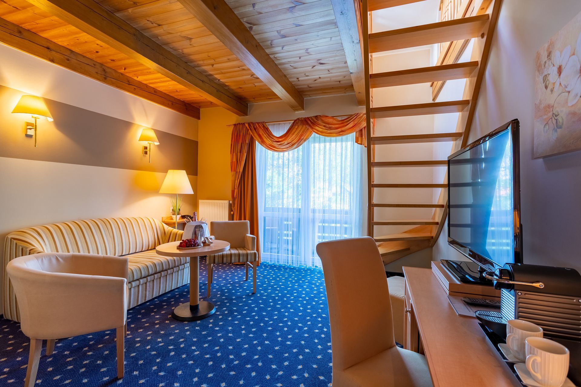 Hotelzimmer mit blauem Teppich, Sofa, kleinem Tisch, Fernseher und Treppe.