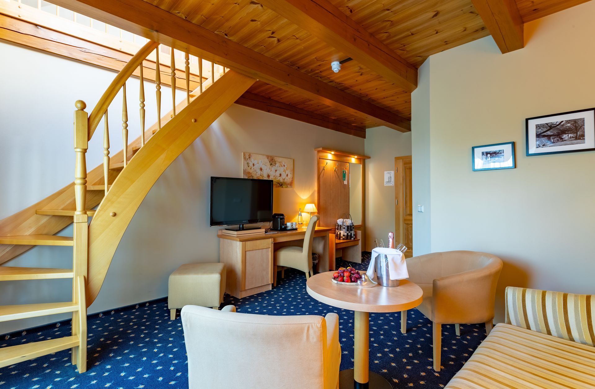 Hotelzimmer mit Holztreppe zu einem Loft, Schreibtisch mit Fernseher, Sitzecke mit kleinem Tisch, blauer Teppich.