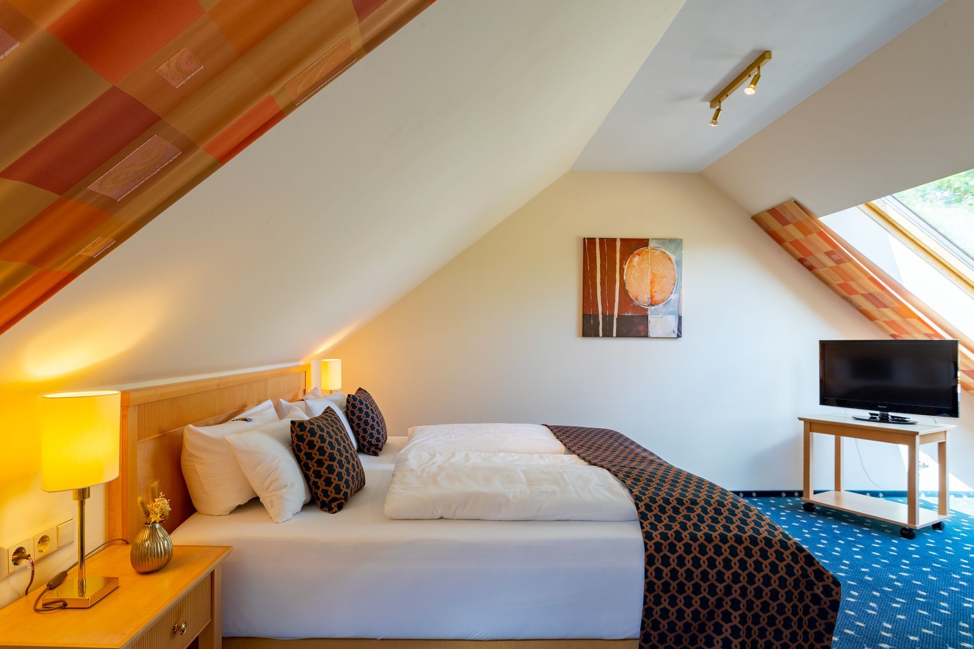 Schlafzimmer mit Bett, Nachttisch, Fernseher und Dachschrägen. Warme Töne, natürliches Licht.