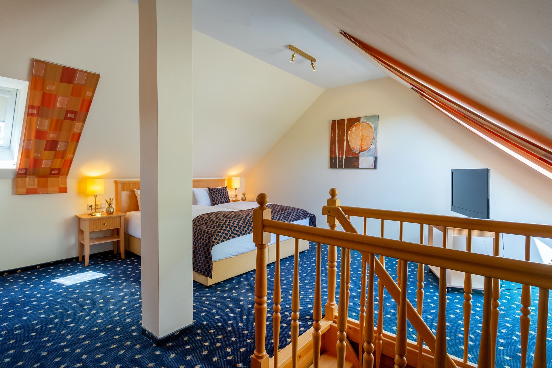 Schlafzimmer mit Bett, Nachttisch, blauem Teppich, Holzgeländer und Dachschräge.