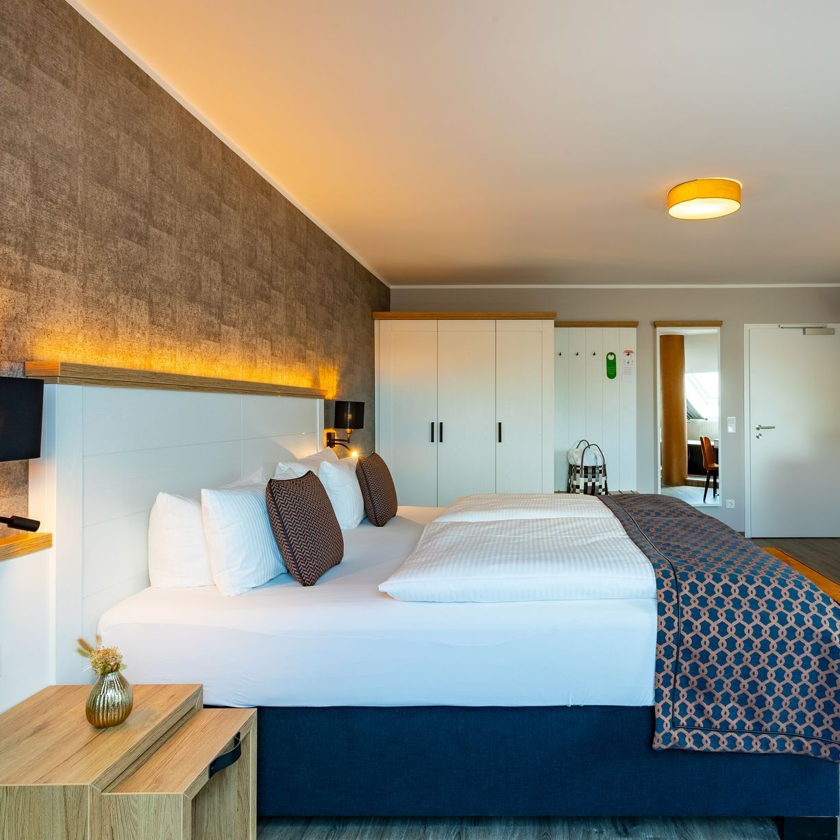 Schlafzimmer mit großem Bett, weißer Bettwäsche, gemusterter Decke, Nachttisch und strukturierter Akzentwand.