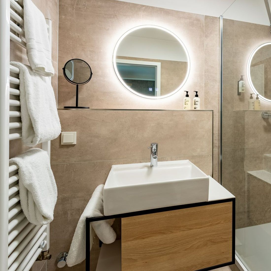 Badezimmer mit weißem Waschbecken, rundem Spiegel mit Beleuchtung und Handtüchern auf einem Heizkörper.