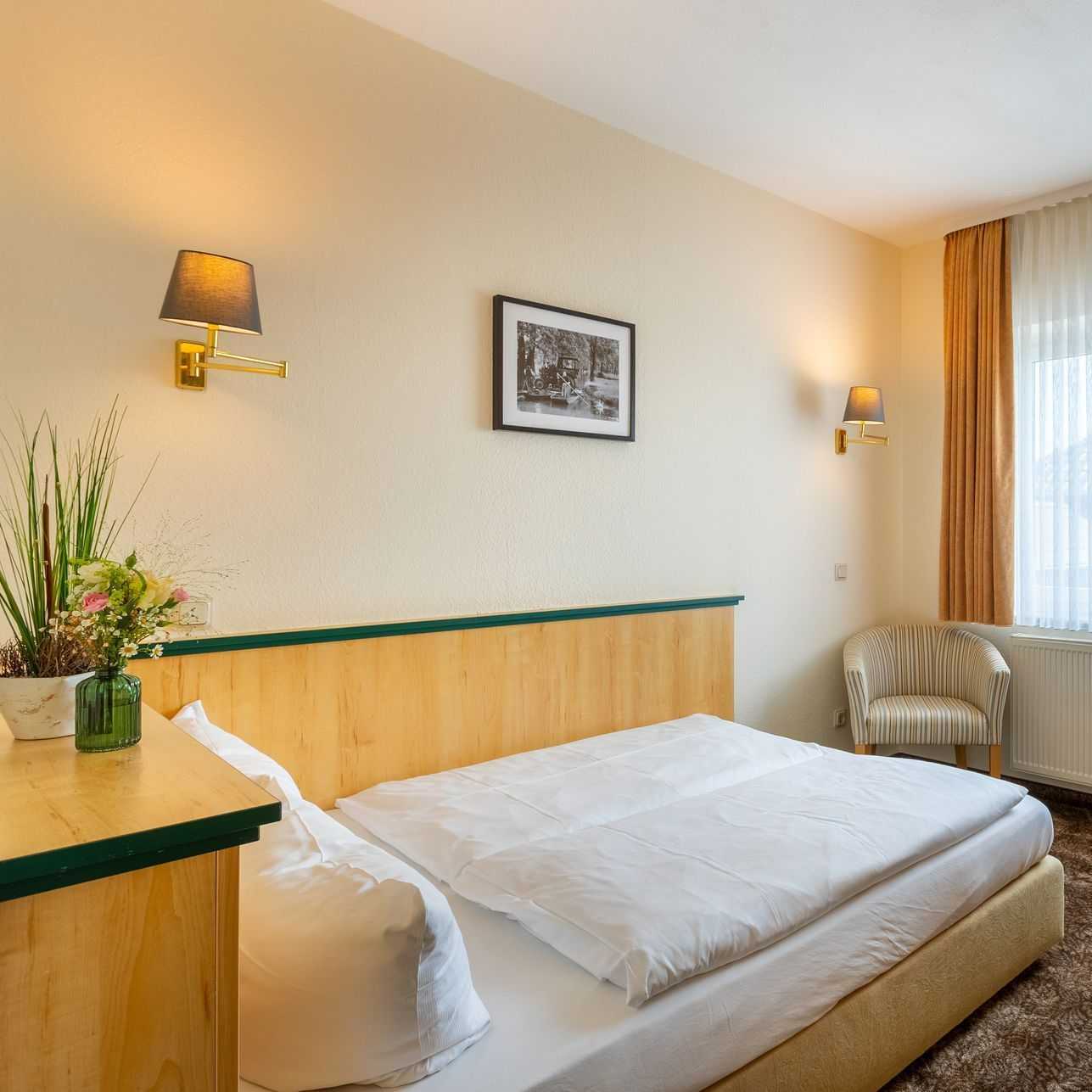 Schlafzimmer mit gemachtem Bett, Beistelltisch mit Blumen, Sessel, Wandleuchten und einem gerahmten Bild.