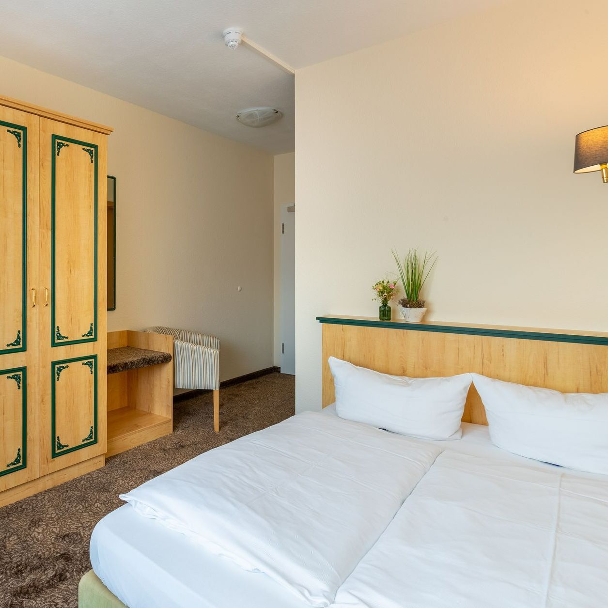 Hotelschlafzimmer mit Doppelbett, Kleiderschrank und Stuhl. Helles Holz und beige Wände.