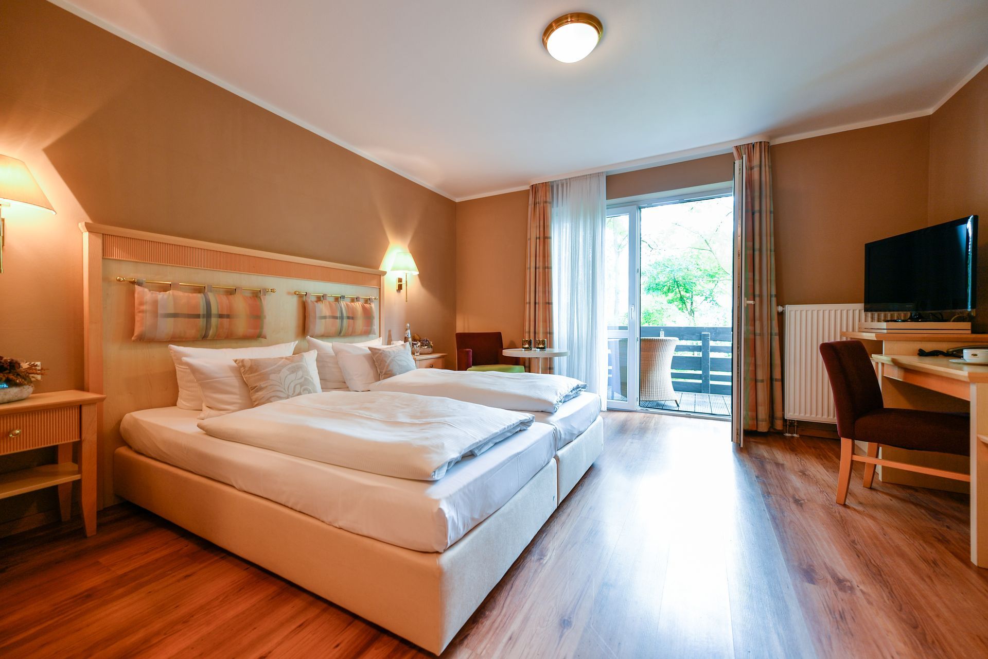 Hotelzimmer mit zwei Betten, Balkon, Schreibtisch und Fernseher. Erdtöne und Holzböden.