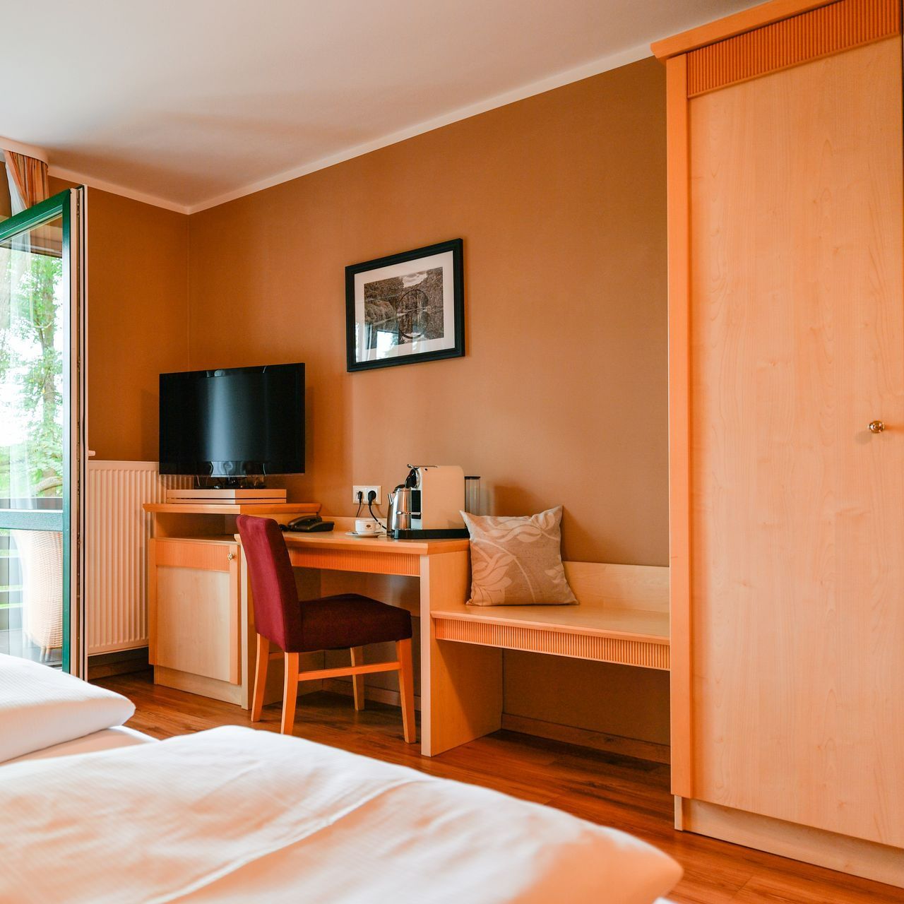 Hotelzimmer mit Bett, Schreibtisch, Fernseher, Schrank. Orange Wände, roter Stuhl, helle Holzmöbel.