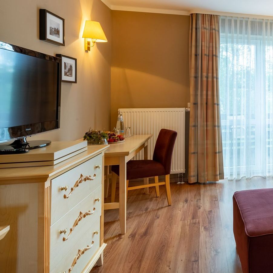 Hotelzimmereinrichtung mit Kommode, Schreibtisch, Stuhl, Fernseher und Fenster mit Vorhängen.