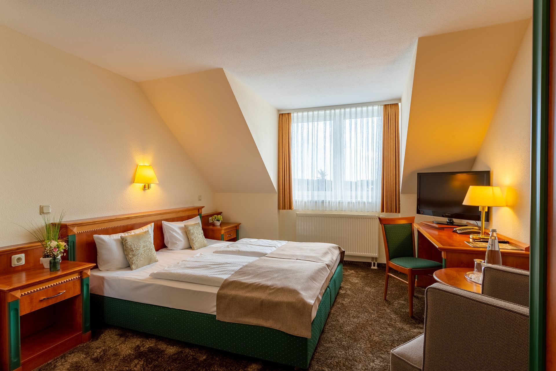Hotelzimmer mit Bett, Fernseher, Schreibtisch und Stuhl. Helle Wände, Teppich und zwei Lampen.
