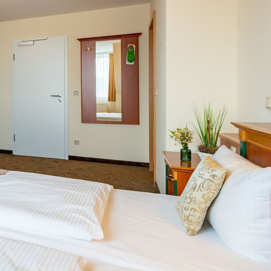 Hotelzimmer: Bett mit weißer Bettwäsche, Spiegel, Tür und Nachttisch mit Blumen.