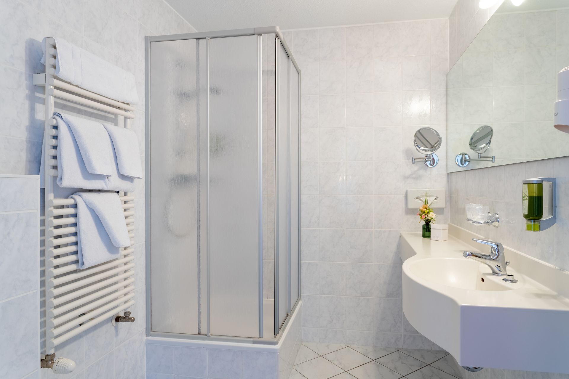 Badezimmer mit Milchglasdusche, weißem Waschbecken, Spiegel, Handtuchhalter und weiß gefliesten Wänden.