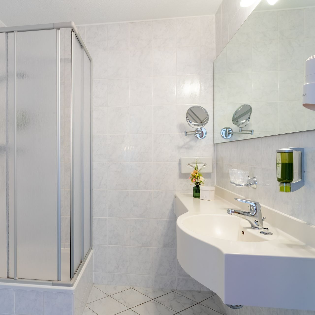 Badezimmer mit weißem Waschbecken, Dusche und Spiegel. Wände und Boden sind weiß gefliest.