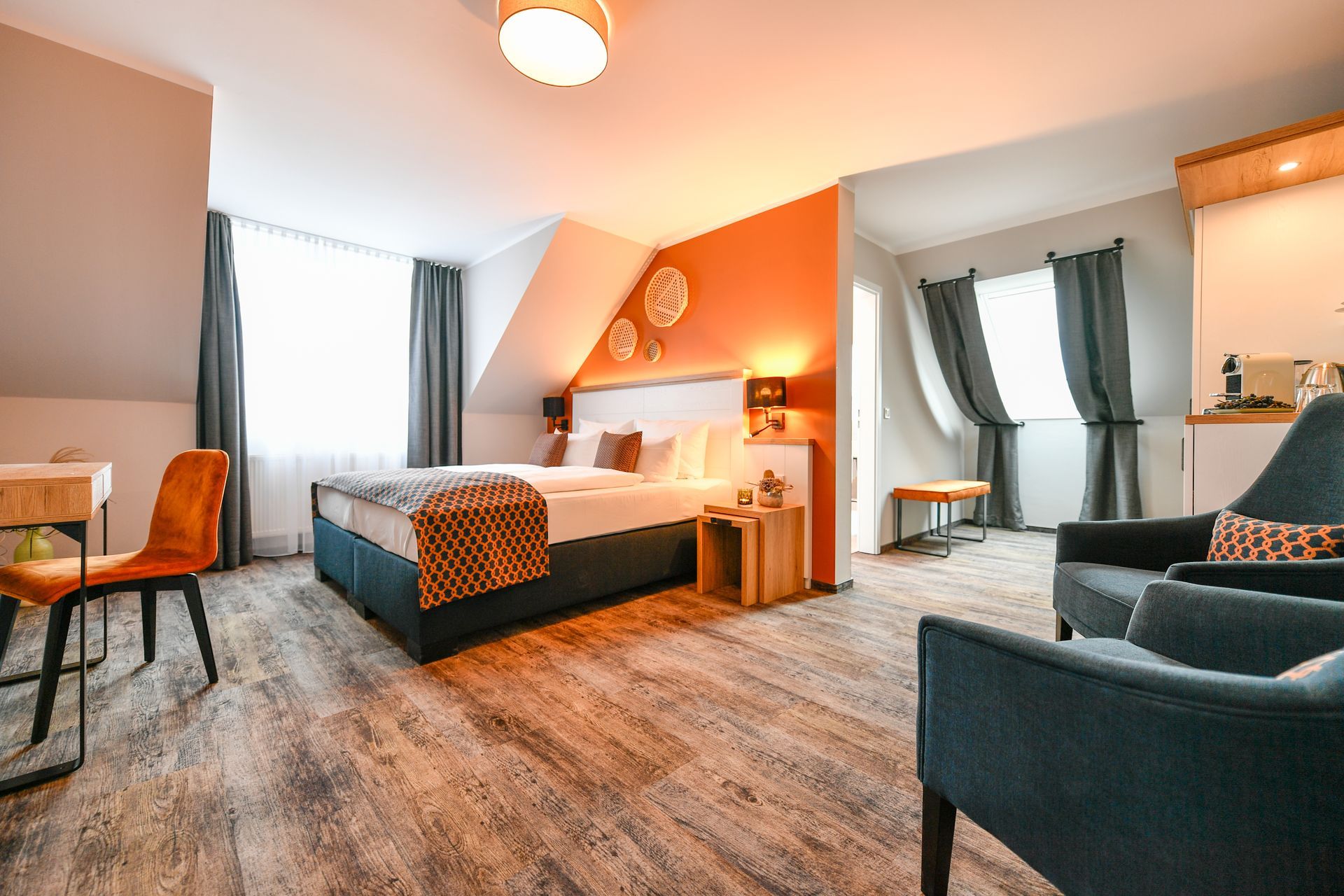 Hotelzimmer mit Bett, Sitzecke, Schreibtisch und Fenster mit Vorhängen; orangefarbene Akzentwand.