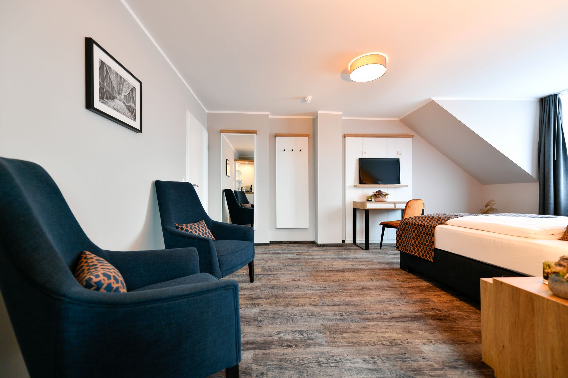 Hotelzimmer mit blauen Sesseln, einem Bett, einem Schreibtisch und einem Fernseher.