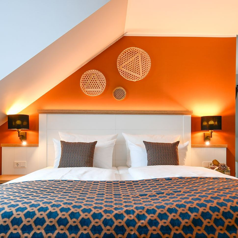 Schlafzimmer mit orangefarbener Akzentwand, weißem Bett und gewebter Wanddekoration.