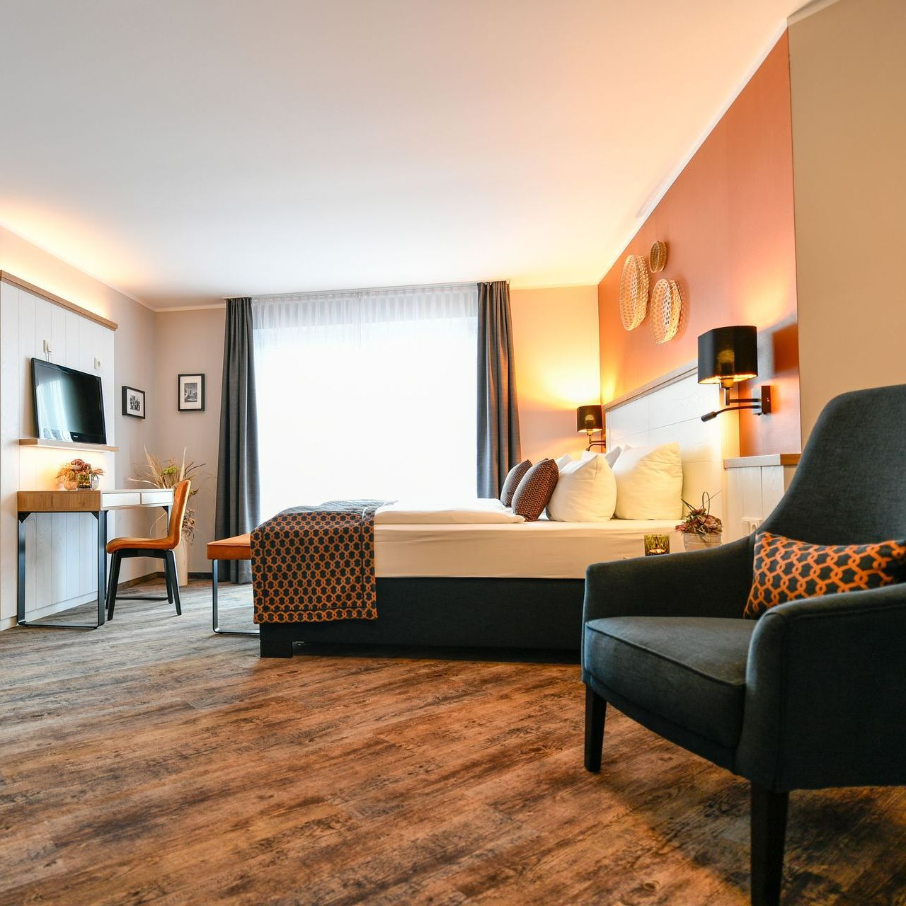 Hotelzimmer mit Bett, Schreibtisch, Sessel und Fenster, in warmen Farben gehalten.