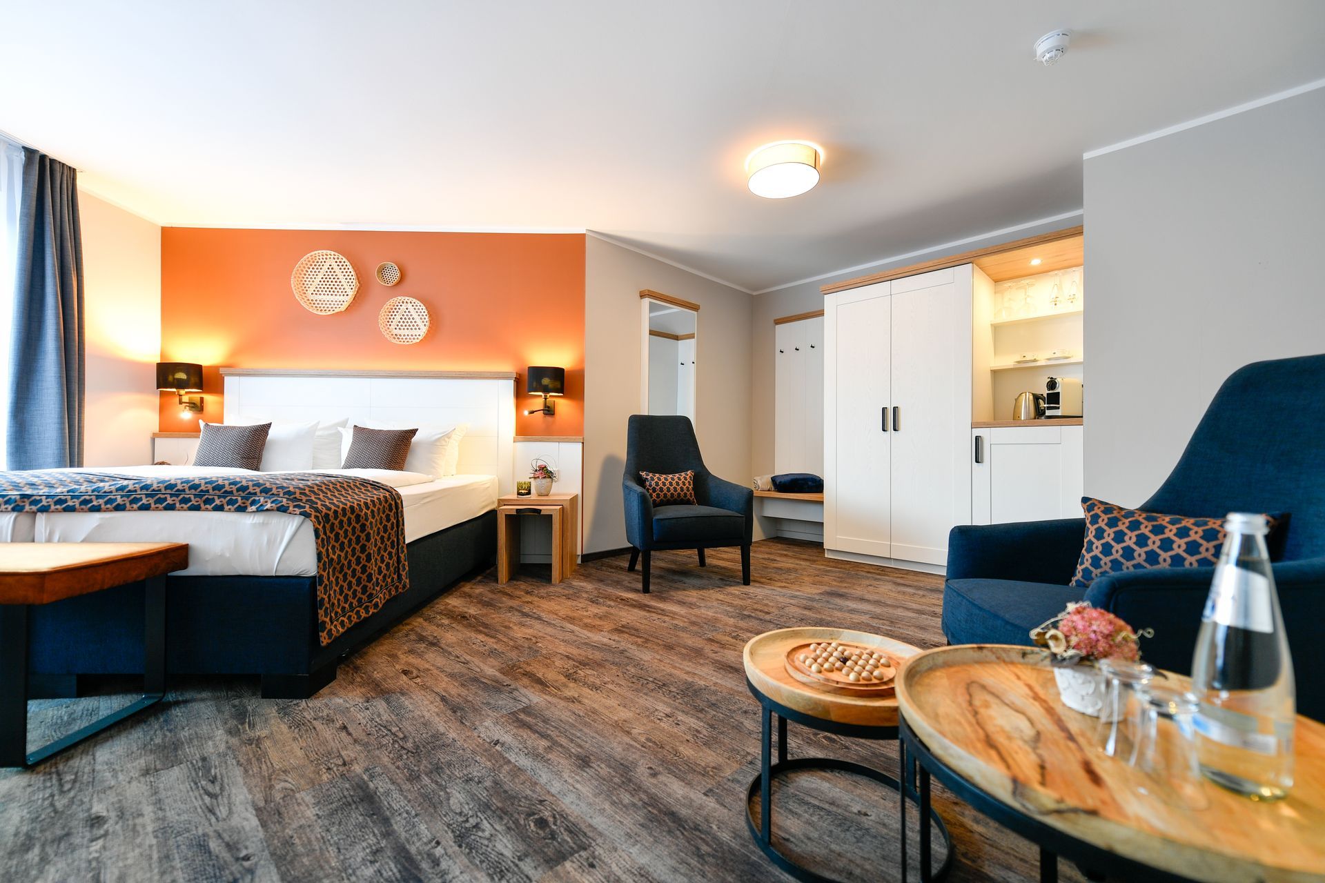 Hotelzimmer mit Bett, Sitzecke mit blauen Stühlen, orangefarbener Akzentwand, Holztischen.