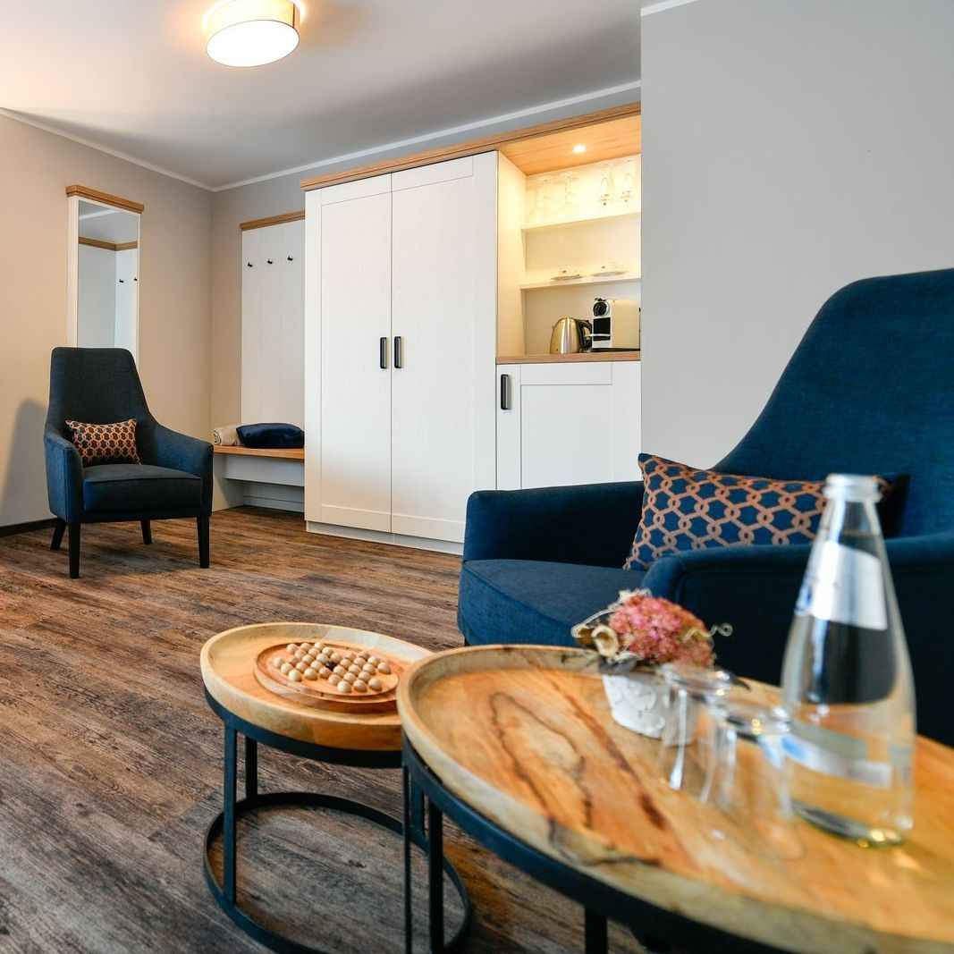 Gemütliches Hotelzimmer mit blauem Sessel, Holztischen und Wasserflasche, neben einer Küchenzeile.