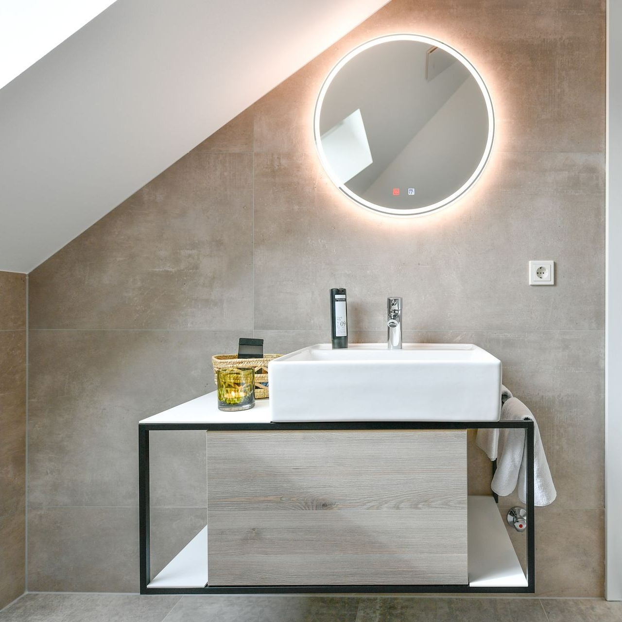 Modernes Badezimmer mit schwebendem Waschbecken, rundem beleuchtetem Spiegel und schräger Decke.