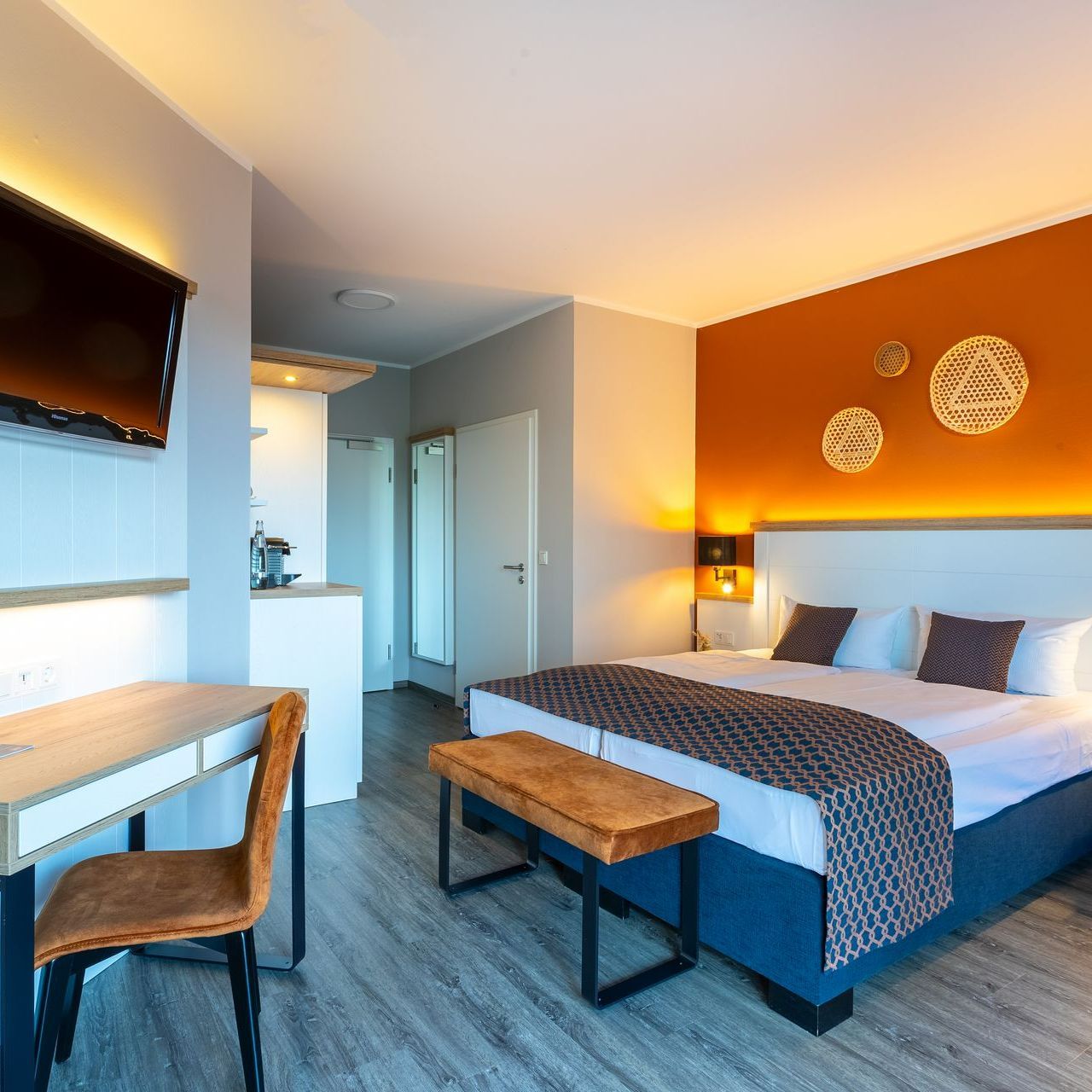 Modernes Hotelzimmer mit orangefarbener Akzentwand, Bett, Schreibtisch und kleiner Küchenzeile.