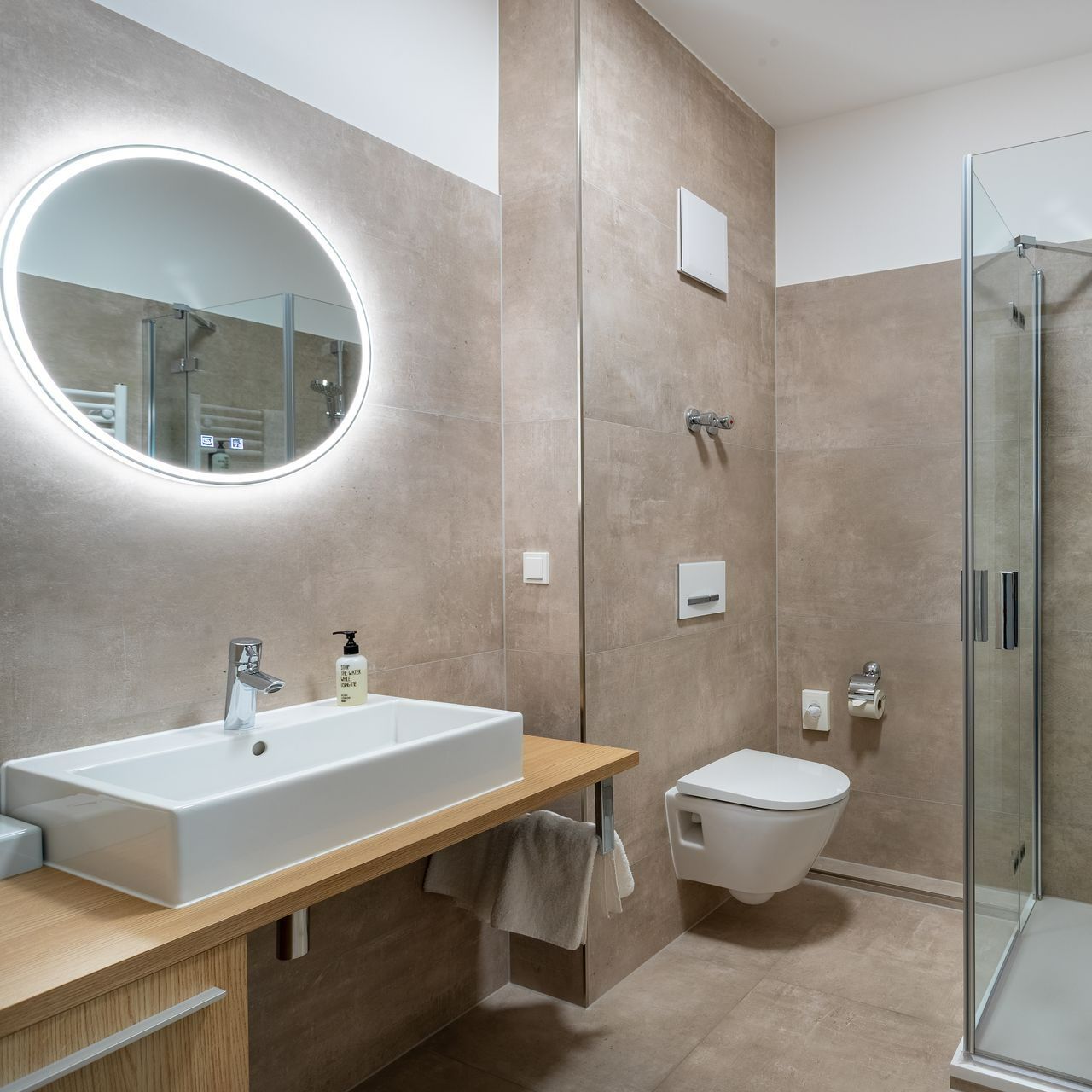 Modernes Badezimmer mit Waschbecken, Toilette, Dusche und rundem Spiegel. Beige Wände und helle Holzakzente.