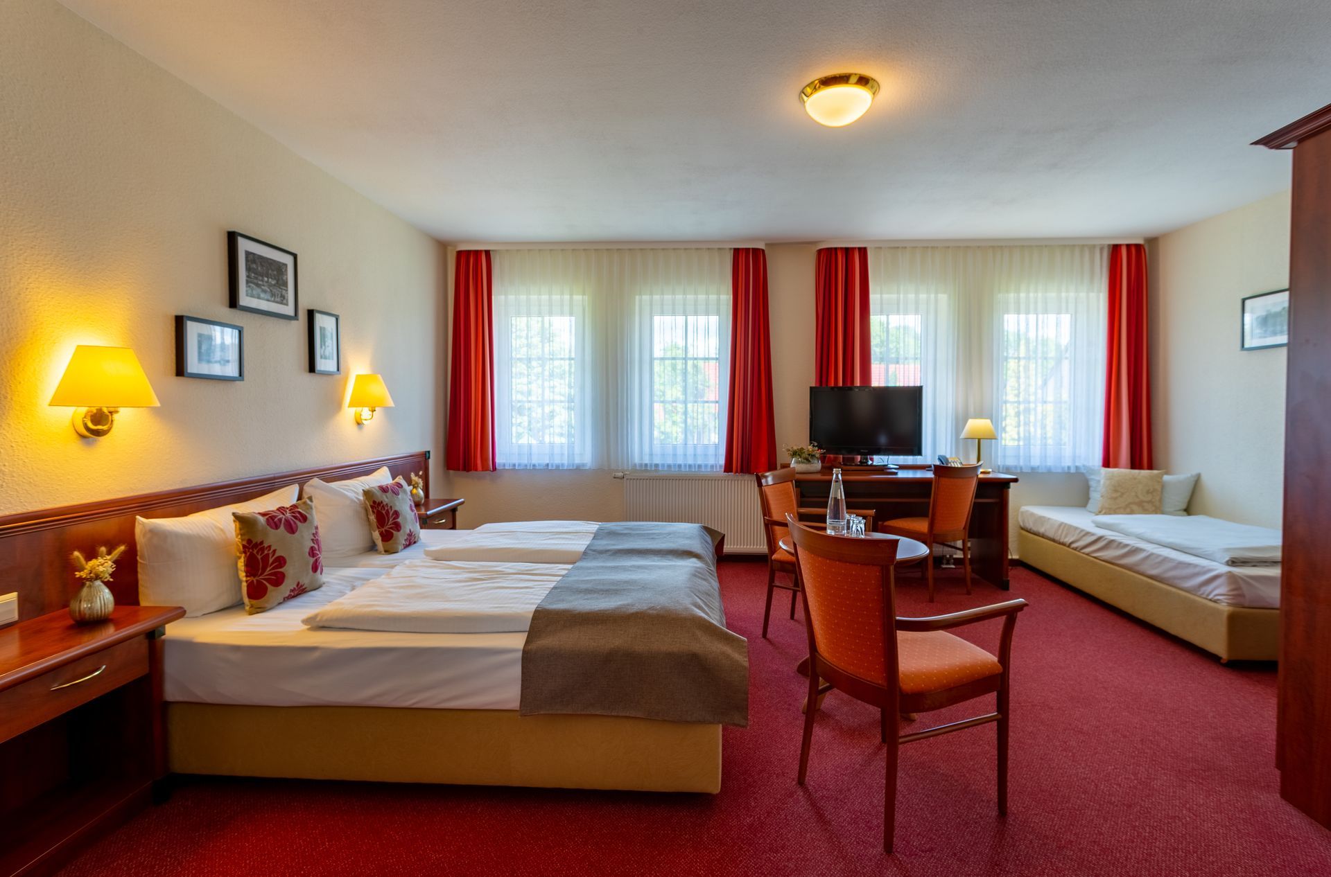 Hotelzimmer mit zwei Betten, einem Sofa, einem Fernseher und roten Akzenten.