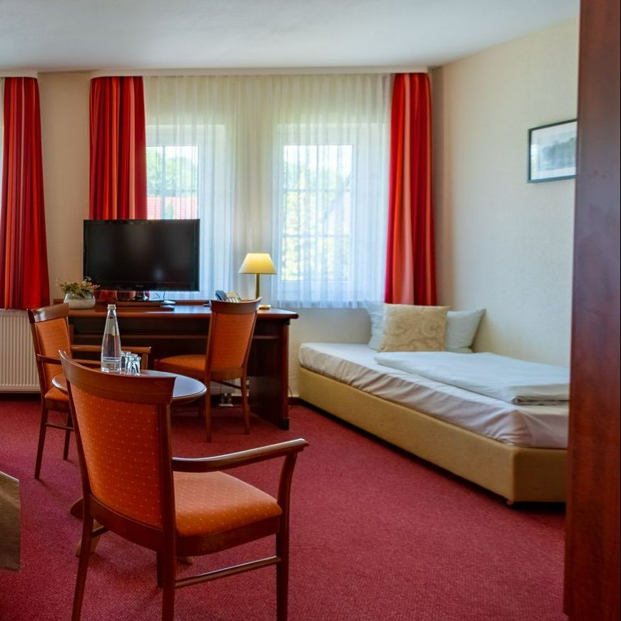 Hotelzimmer mit rotem Teppich, Schreibtisch, Bett, Fernseher, roten Vorhängen und zwei orangefarbenen Stühlen.